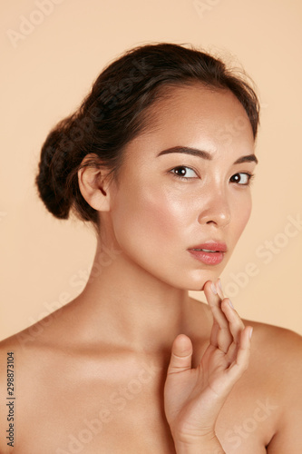 Beauty face. Woman with natural makeup and healthy skin portrait. Beautiful asian girl model touching fresh glowing hydrated facial skin on beige background closeup. Skin care concept