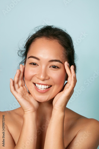 Beauty face. Smiling asian woman touching healthy skin portrait. Beautiful happy girl model with fresh glowing hydrated facial skin and natural makeup on blue background at studio. Skin care concept