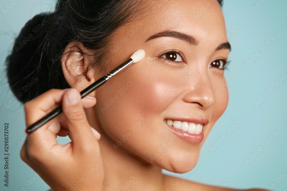 Beauty. Woman applying makeup on eyes with cosmetic brush closeup. Portrait of happy smiling asian girl model with beautiful face applying facial make up at studio