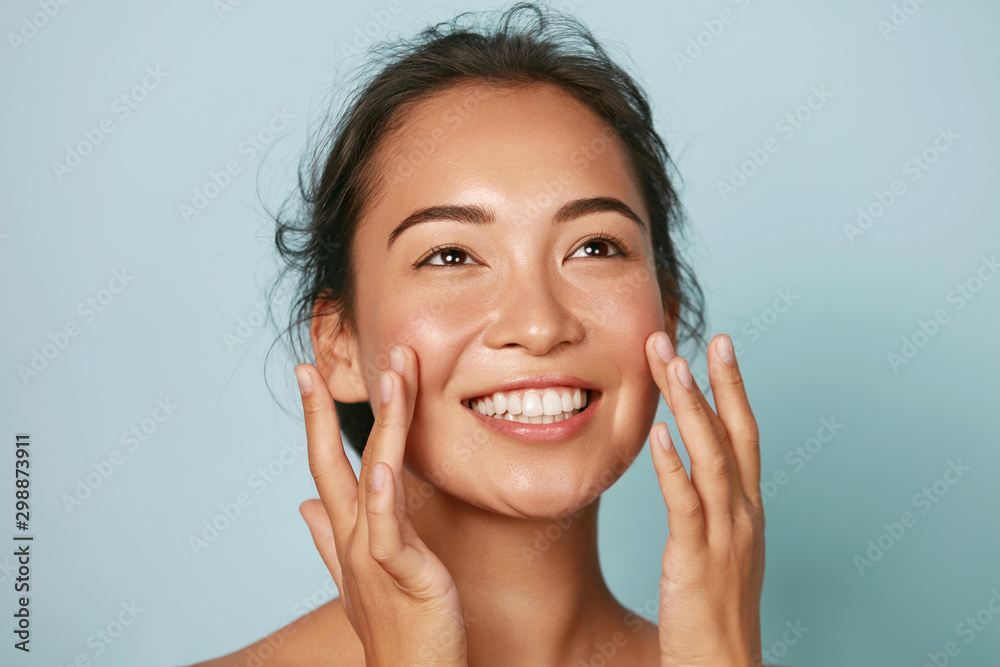 Beauty face. Smiling asian woman touching healthy skin portrait. Beautiful happy girl model with fresh glowing hydrated facial skin and natural makeup on blue background at studio. Skin care concept - obrazy, fototapety, plakaty 