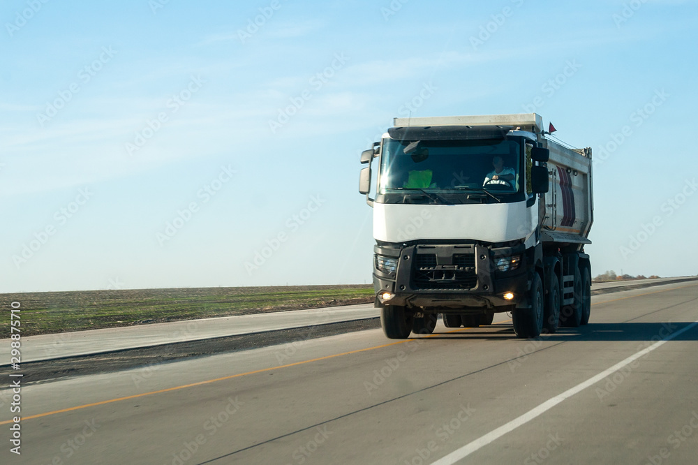 A white dump truck drives out of town on a flat asphalt road among fields against a blue sky on a sunny day. A machine for transporting building materials rides on the highway