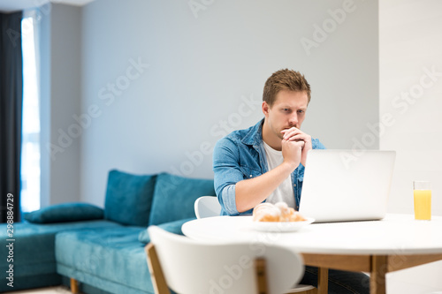 young handsome man busy in his laptop while having croissant and fresh orange juice for lunch working from home looking serious