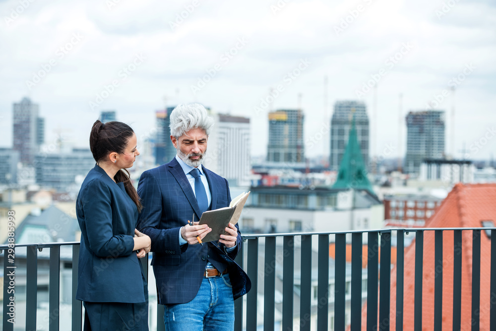 A businessman and businesswoman with diary standing on a terrace, working. A businessman and businesswoman with diary standing on a terrace, working.