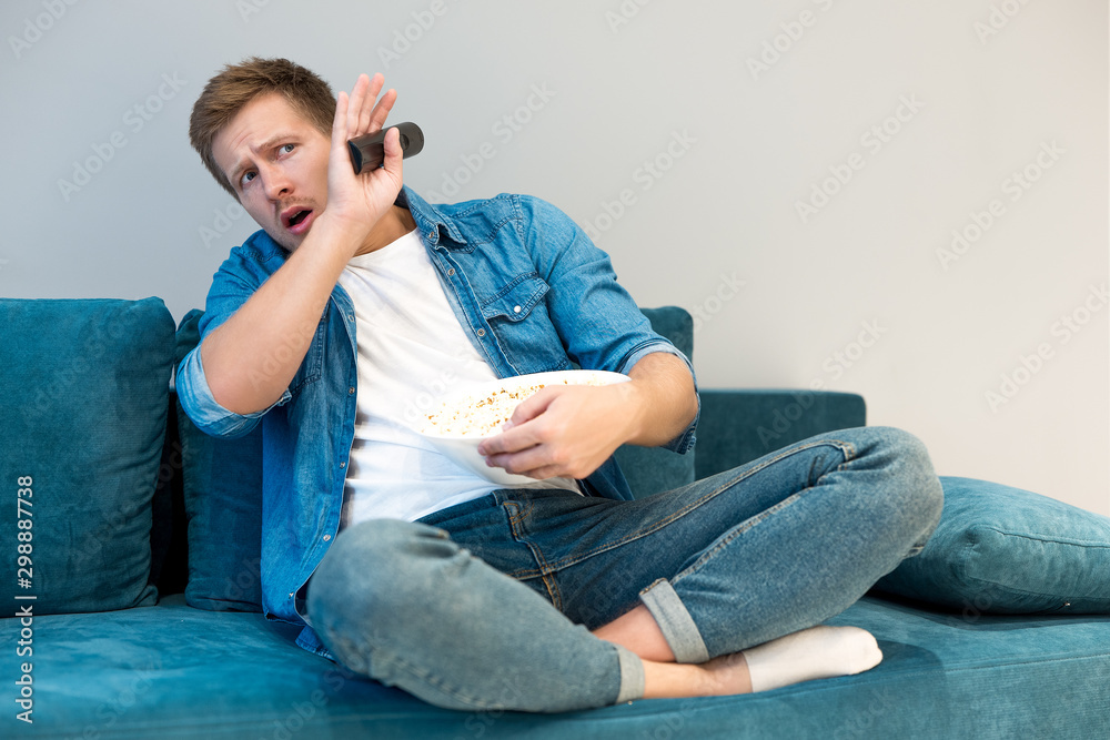 Fototapeta premium young handsome frightened man watching scary movie on the sofa eating popcorn feeling comfortable at home