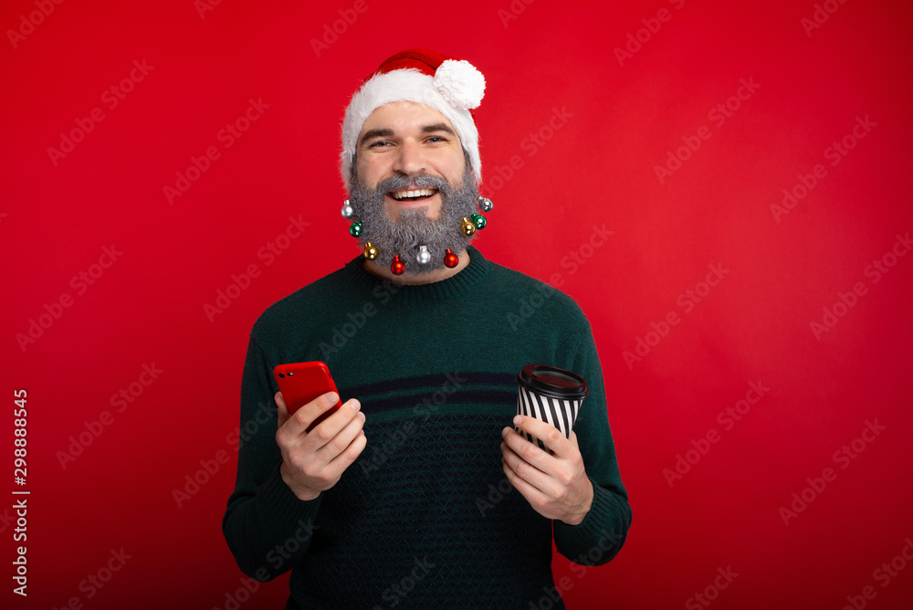 Happy bearded man wearing santa claus hat holding smartphone and cup of coffee to go Happy bearded man wearing santa claus hat holding smartphone and cup of coffee to go