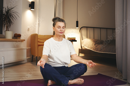 Senior woman in sportswear sitting barefooted on floor at home, keeping legs crossed on mat, doing meditation after yoga practice, having calm peaceful facial expression. Retirement and well being