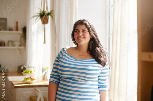 Beauty, bodypositivity, people and lifestyle concept. Indoor shot of gorgeous brunette Latin girl posing by window in kitchen dressed in xxl blue and white striped shirt, having joyful beaming smile photo