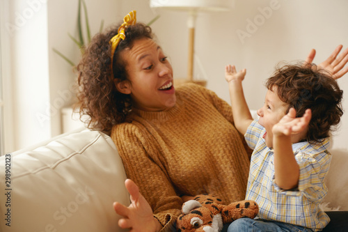 Attractive cheerful young Latin mother telling fairytale to her baby son having excited looks, waving hands and laughing. Happy mom spending maternity leave playing with her adorable baby at home