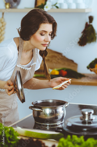 Young brunette woman cooking soup in kitchen. Housewife holding wooden spoon in her hand. Food and health concept