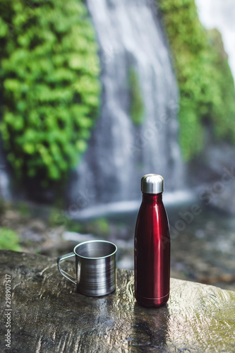 Red thermos bottle and steel cup with background of tropical waterfall. Zero waste eco concept. Hiking equipment.