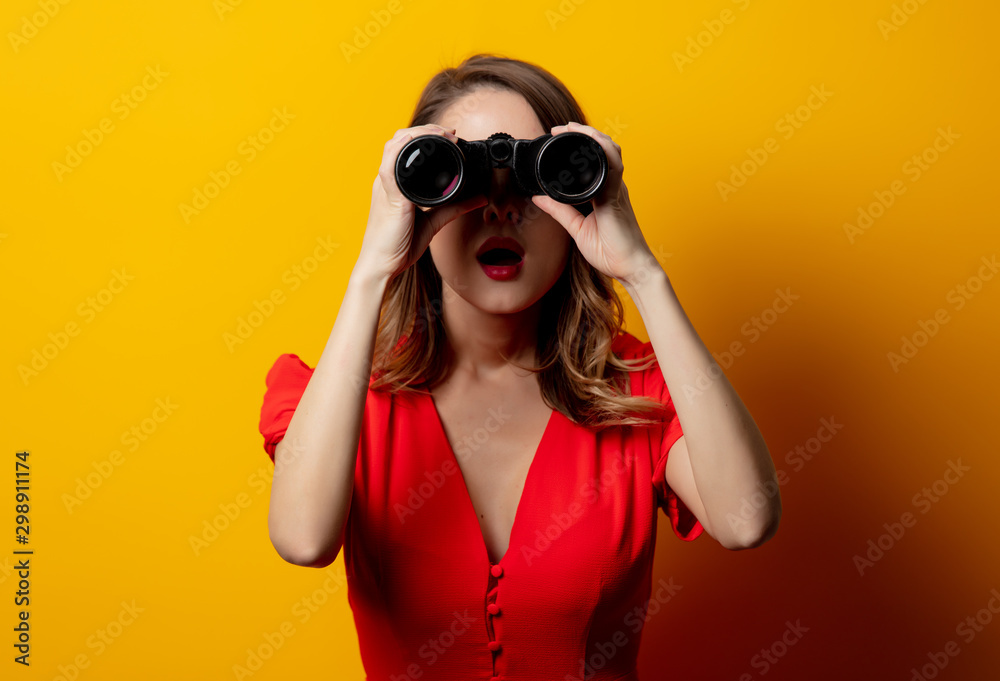 Young woman in red dress with binocular on yellow background Young woman in red dress with binocular on yellow background