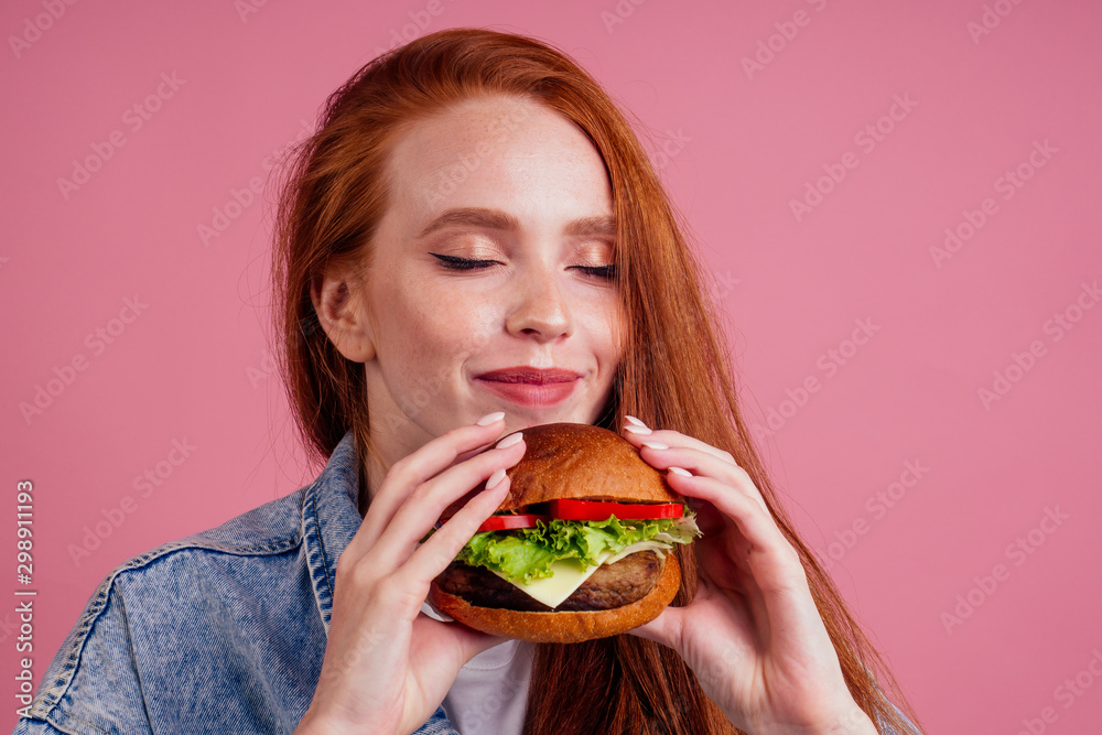 redhaired ginger woman with freckles enjoying big huge burger cutlet and wearing demin american jeans jacket in studio pink background .USA traditional concept redhaired ginger woman with freckles enjoying big huge burger cutlet and wearing demin american jeans jacket in studio pink background .USA traditional concept