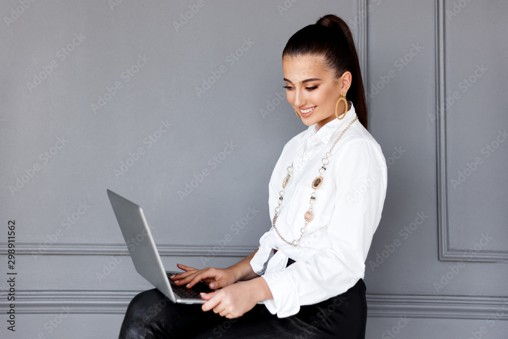 Confident young business woman with perfect make-up, smiling and looking at the laptop, isolated on a grey background. Job, working modern concept. Copy space. Confident young business woman with perfect make-up, smiling and looking at the laptop, isolated on a grey background. Job, working modern concept. Copy space.
