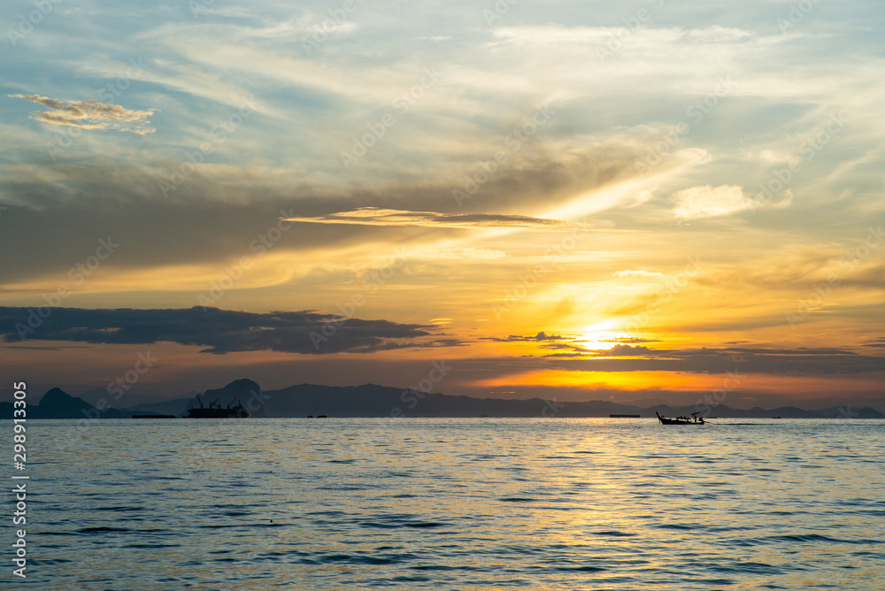 Fototapeta premium Silhouette of moving long tail boat with shadow of island and orange light of sunrise in background. Landscape of calm sea in early morning