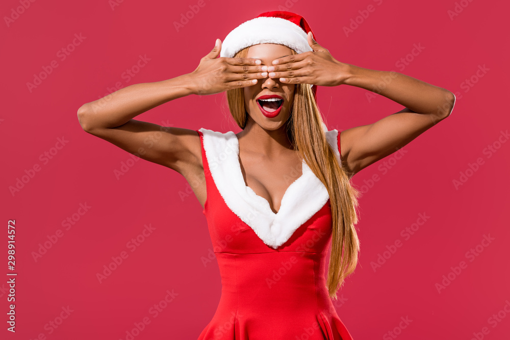 cheerful african american girl in santa hat and christmas dress covering eyes with hands isolated on red cheerful african american girl in santa hat and christmas dress covering eyes with hands isolated on red