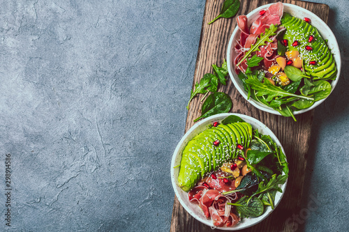 Low carbs bowl. Fresh salad with green spinach, rucola, avocado an ham serrano in white bowl, gray background, top view photo