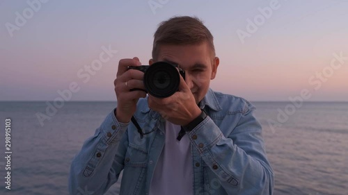 A male photographer on the beach looks into the camera and takes a photo. Sea in the background. Slow motion.