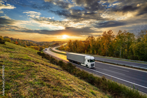 Obraz na płótnie White trucks driving on the asphalt highway in autumn landscape at golden sunset with dramatic clouds w salonie