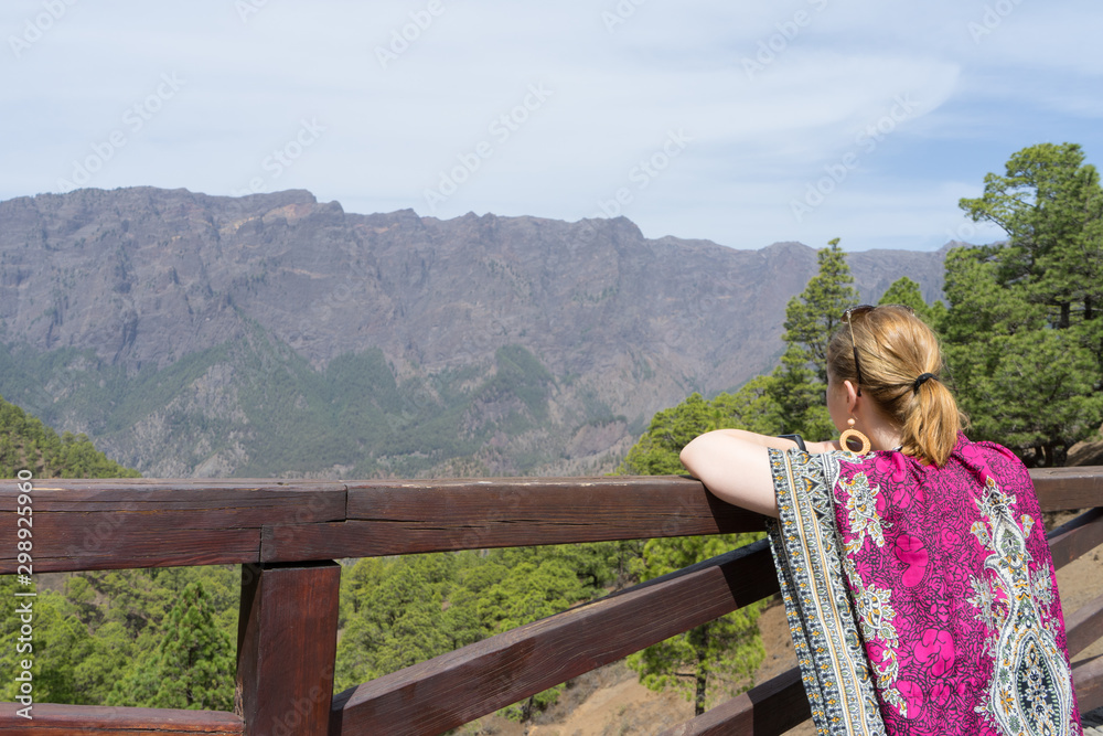 Naklejka premium Teen tourist resting, hold a wood fence, relaxing and look the environment national park with colorful dress. Blonde woman observe and enjoy the forest landscape in a view point.