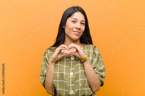young pretty hispanic woman smiling and feeling happy, cute, romantic and in love, making heart shape with both hands against brown wall photo