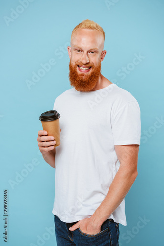 smiling bearded man in t-shirt holding coffee to go, isolated on blue