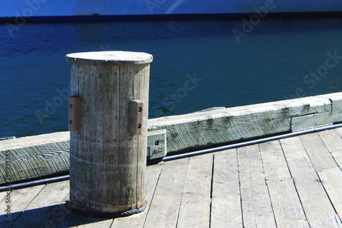 Wooden bollard on a wooden pier, harbour side. Partial view of water and a ship at its waterline.