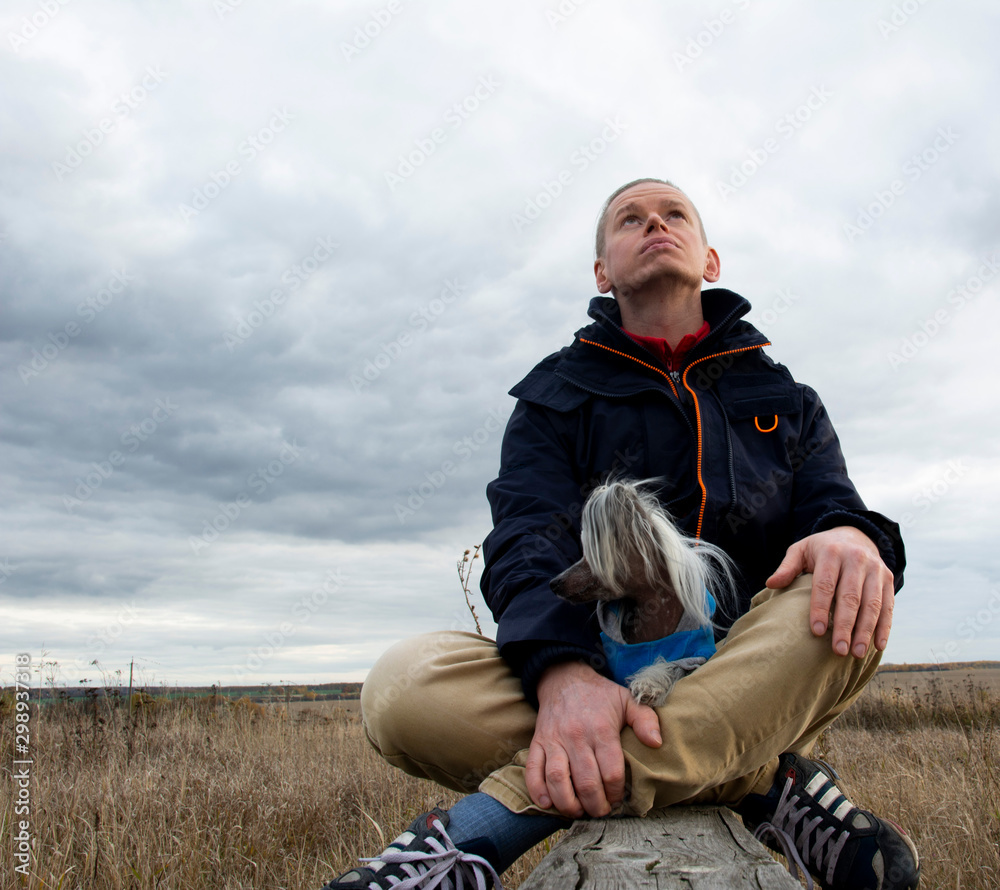 A middle-aged man dreams while looking at the sky on an autumn day in nature. Nearby is a dog breed Chinese Hairless Crested Dog. A middle-aged man dreams while looking at the sky on an autumn day in nature. Nearby is a dog breed Chinese Hairless Crested Dog.