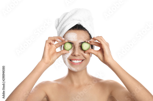 Happy young woman with organic mask on her face holding cucumber slices against white background