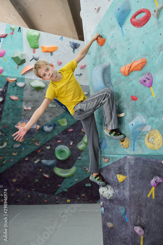 Cute schoolboy in activewear holding with one hand by small rock on climbing wal