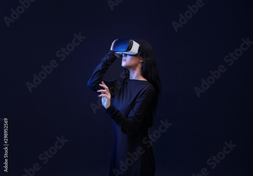 Woman in VR helmet scrolling invisible screen while interacting with virtual reality under neon light. Girl in glasses of virtual reality. Augmented reality, game concept. Dark background.