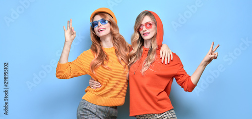 Two Lovable embracing fashionable woman sisters dance in Trendy orange outfit. Studio shot of Carefree funny stylish friends laughing on blue. Happy fashion girl, dancing positive mood, peace sign © evgenij918