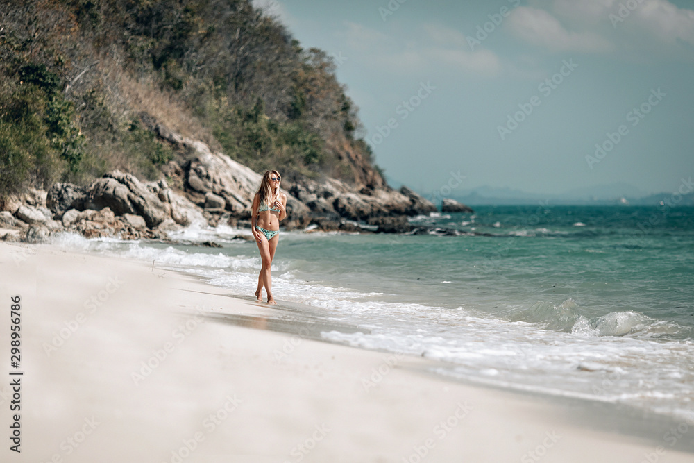 Attractive slim blondie in bikini walks along the sandy beach against the blue bay, sunny day, relaxation.