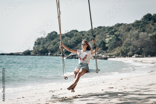 Attractive slim girl on the rope swings, sandy beach against the blue bay, sunny day, relaxation.