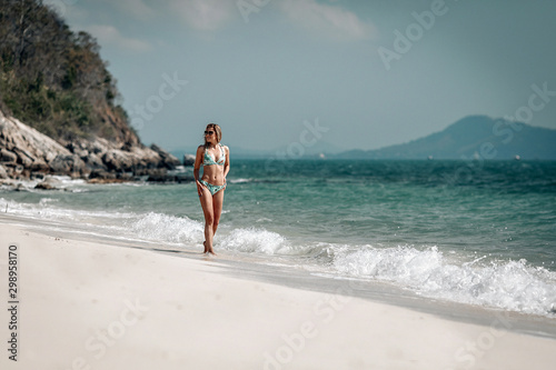 Attractive slim blondie in bikini walks along the sandy beach against the blue bay, sunny day, relaxation.