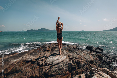.Woman in black swim suit on a cliff by the ocean in a sunny day, her arms above her head, back side view, silhouette..