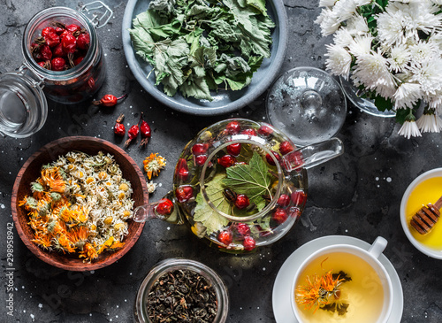 Ingredients for herbal tea - currant, mint, raspberry leaves, chamomile and calendula flowers, rose hips and teapot on a dark background, top view