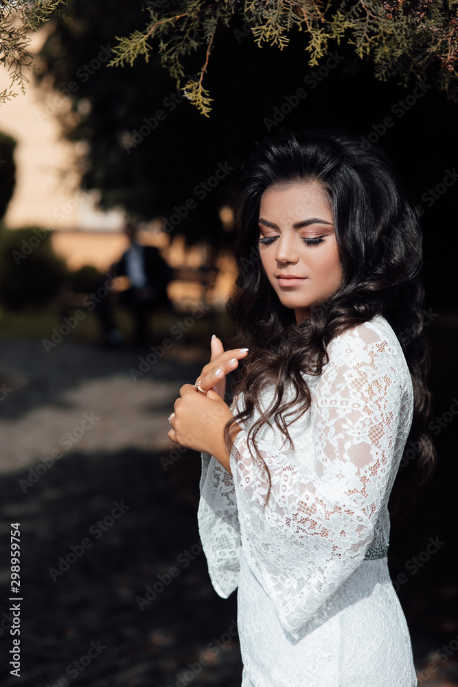 Naklejka premium Cute smiling woman with long wavy hair and makeup, attractive young girl enjoying summer in luxury white dress. Portrait of young woman standing on the park on sunny summer or spring day outside.