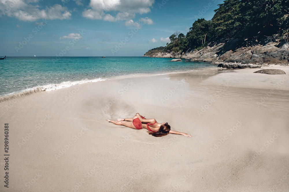Sexy girl in red bikini sunbathing on wet sand by the clear ocean, sunny day, rocks, trees; sunbathing concept.