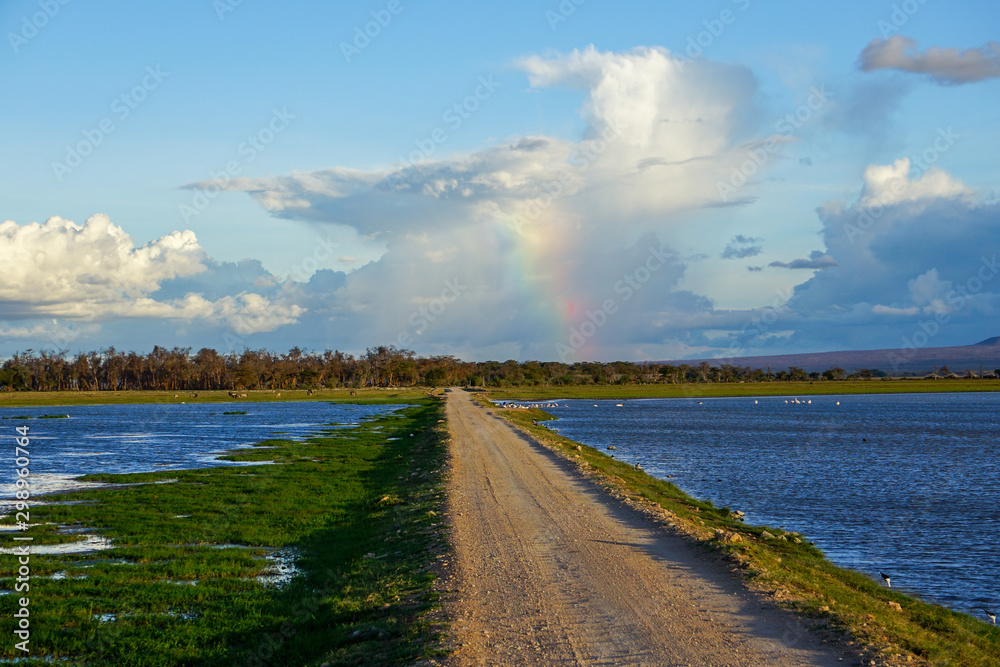 View of sandy safari road running through lake with rainbow in the distance. Bright day, blue sky with soft white clouds. Amboseli National Park -Kenya