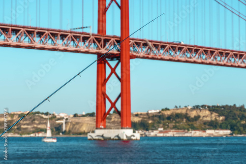 fishing pole against red bridge on the bank of Tagus river. Suspension Bridge over river on background.