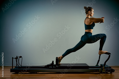 Young girl doing pilates exercises with a reformer bed. Beautiful slim fitness trainer on a reformer gray background, low key, art light, copy space advertising banner photo