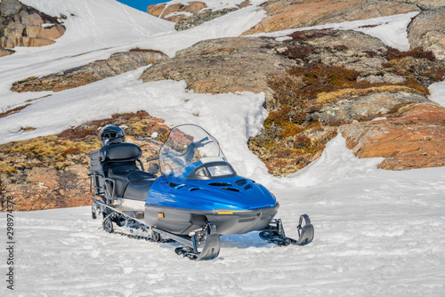 One blue snowmobile stands on snow close to stones in Norwegian mountains, warm day and bright sun, snow smelts, norwegian mountains landscape, front side view, black safety helmet lays at back side photo