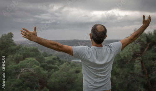 An aged happy man standing on top of a mountain with his arms outstretched. Beautiful view of the mountains and sky from the back of a man
