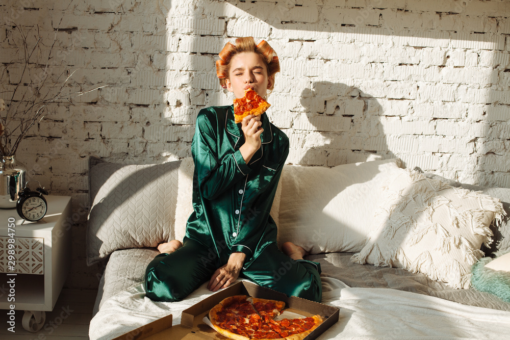 Φωτογραφία attractive young woman in green pajamas and hair curlers eating pizza in bed