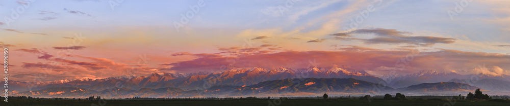 Panorama. Fairytale dawn, sunset with bright multi-colored clouds, against the backdrop of a mountain range with snow-capped peaks