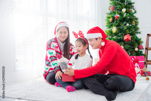 Young Asian girl opening Christmas present with her father and mother In a room decorated with Christmas trees.