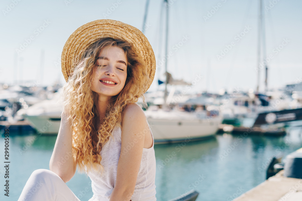 curly blonde sits on a parapet by the sea, closing her eyes with pleasure against the background of ships