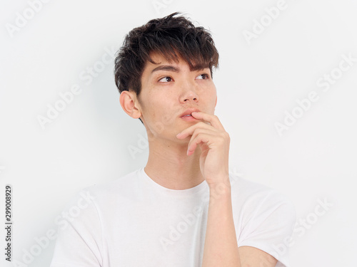 Portrait of a handsome Chinese young man in white t shirt looking away with hand on his lips  thinking expression  isolated on white background. 