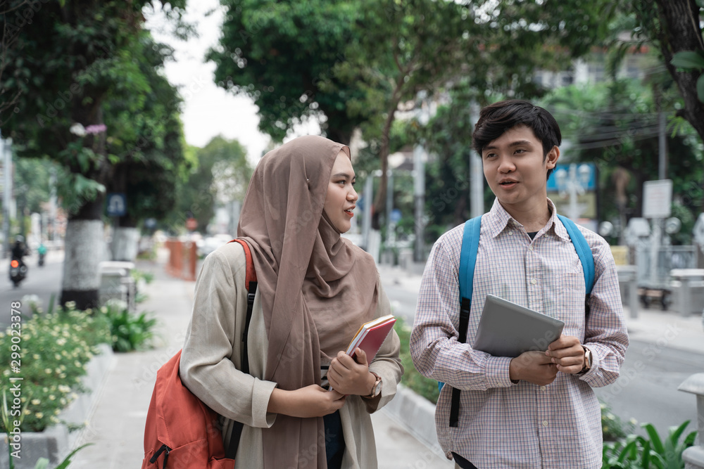 male and female student holding tablet and books walking in the park