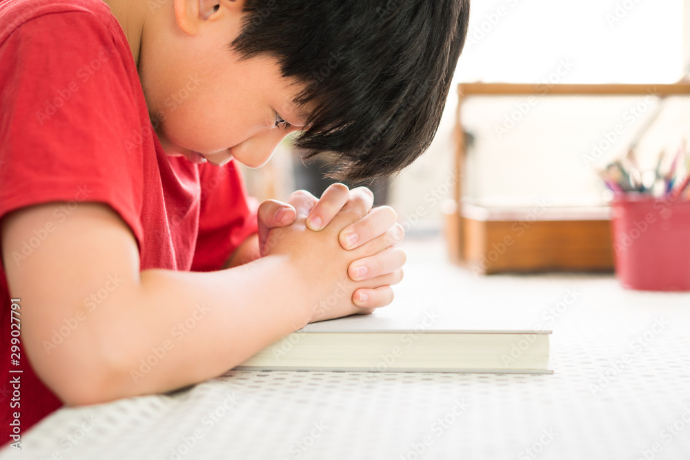 Closeup : Smart looking teen Asian boy desperately praying with faith to God with opened eyes looking at his fingers, almost cry, joined hands tight over white hard cover bible in his bedroom. Closeup : Smart looking teen Asian boy desperately praying with faith to God with opened eyes looking at his fingers, almost cry, joined hands tight over white hard cover bible in his bedroom.